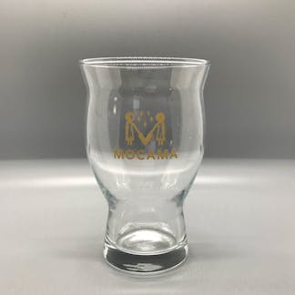 This is a picture of a glass with a slim base and a wide, flared out top. It has a printed logo in gold which contains the words Mocama and an illustration of two women holding barley stalks.