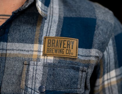 Blue and grey plaid flannel shirt with leather Bravery Brewing logo patch above left chest pocket