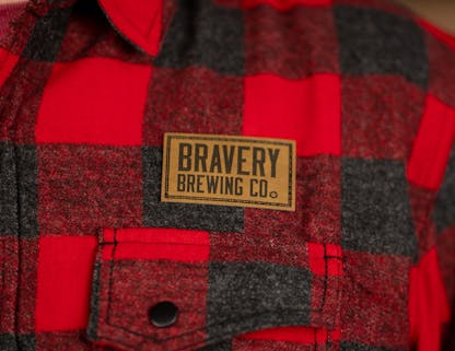 Red and black plaid flannel shirt with leather Bravery Brewing logo patch above left chest pocket
