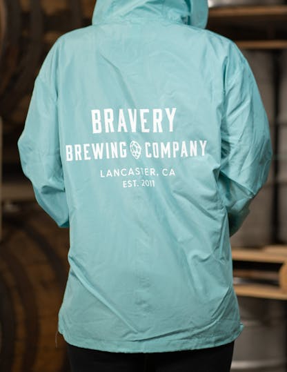 light blue windbreaker jacket with large white text on the back reading, "Bravery Brewing Company, Lancaster CA, est. 2011"
