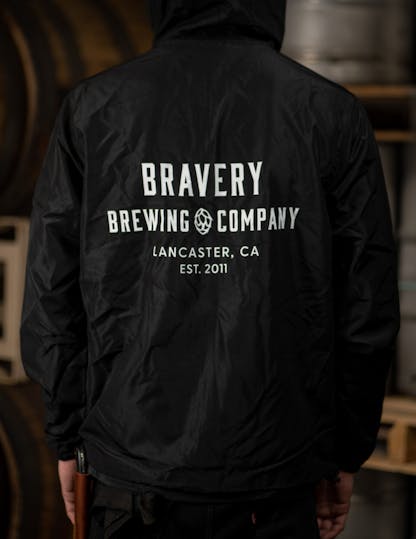 black windbreaker jacket with large white text on the back reading, "Bravery Brewing Company, Lancaster CA, est. 2011"