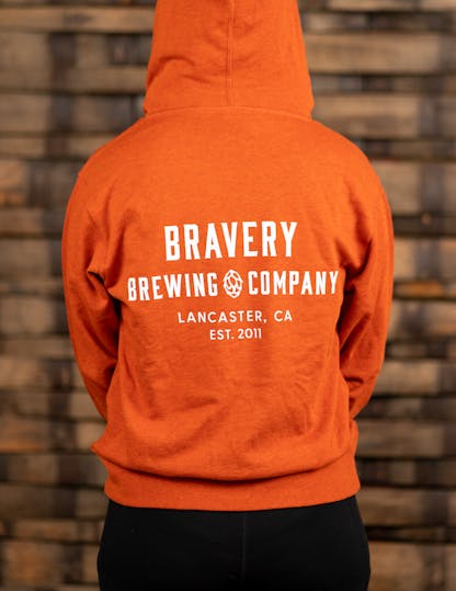 Orange zip-up hoodie with large white text on the back reading, "Bravery Brewing Company, Lancaster CA, est. 2011"