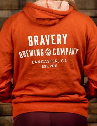 Orange zip-up hoodie with large white text on the back reading, "Bravery Brewing Company, Lancaster CA, est. 2011"