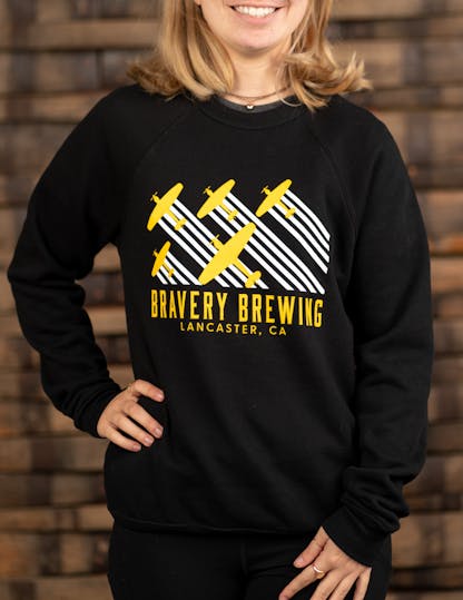Woman in black sweatshirt with yellow and white plane design, "Bravery Brewing, Lancaster, CA"