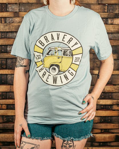 front of light blue t-shirt with a circular design with the text "Bravery Brewing, Est. 2011." In the center of the circle is an illustrated vintage style van with a basset hound sticking it's head out of the car window. The dogs ears are drawn to be flowing in the wind.