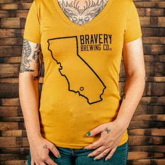 front of a mustard yellow women's V-Neck style t-shirt. There is a large outline of the state of California with a star near the bottom of the state where Bravery is located. Next to the outline of the state is the text "Bravery Brewing Co." Both the illustration and text are printed in black ink.