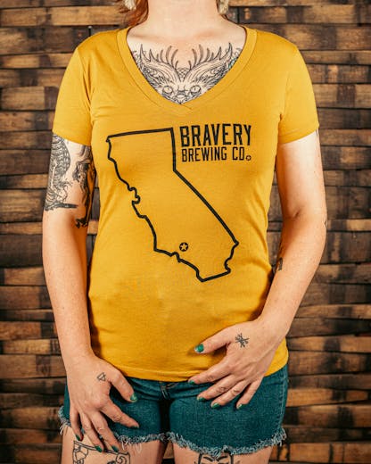 front of a mustard yellow women's V-Neck style t-shirt. There is a large outline of the state of California with a star near the bottom of the state where Bravery is located. Next to the outline of the state is the text "Bravery Brewing Co." Both the illustration and text are printed in black ink.