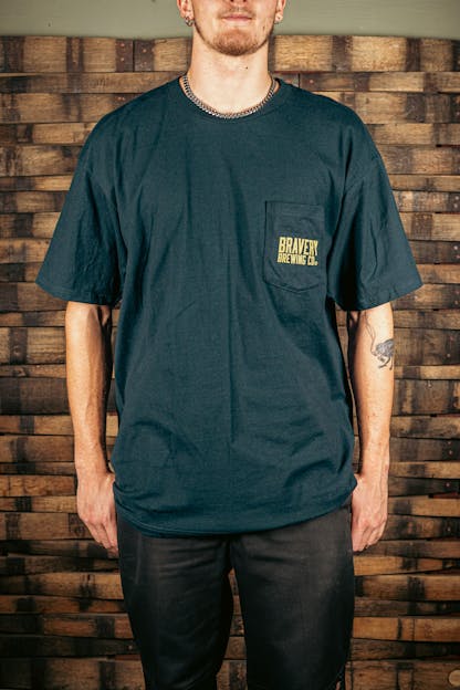 front of a navy t-shirt with a pocket on the wearer's left. printed on the pocked is the text "Bravery Brewing Co." in yellow ink.