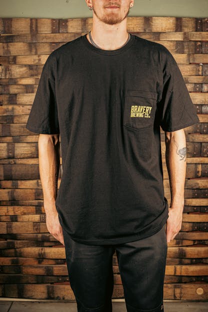 front of a black t-shirt with a pocket on the wearer's left. printed on the pocked is the text "Bravery Brewing Co." in yellow ink.