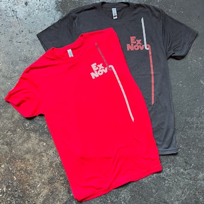 two t-shirts on concrete floor, red partially atop graphite black, both with two-tone front left chest custom logos text "Ex Novo"