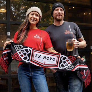 man and woman stand together holding beers and black, red and white scarf with text "Ex Novo Brewing Company" and "Drink Beer Do Good". Woman wears cream colored beanie and red t-shirt with text "Ex Novo". man wears black beanie and t-shirt with text "Ex Novo"