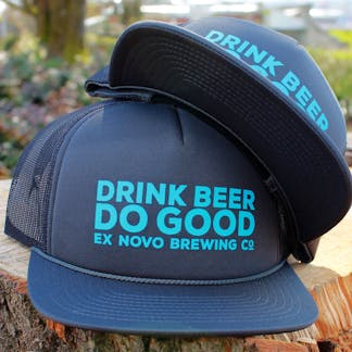 two gray foam baseball hats with mesh backs stacked on a log with teal bold text "Drink Beer Do Good" and smaller teal text "Ex Novo Brewing Co."