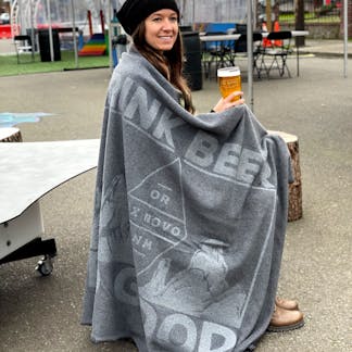 smiling woman with long brown hair wearing black beanie and brown boots sits holding a beer and wrapped in a two tone gray blanket with mountains and large text "Drink Beer Do Good" and within diamond logo text "OR - Ex Novo - NM"