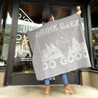 woman wearing black beanie and brown boots holds up beer and two tone gray blanket with mountains and large text "Drink Beer Do Good" and within diamond logo text "OR - Ex Novo - NM"