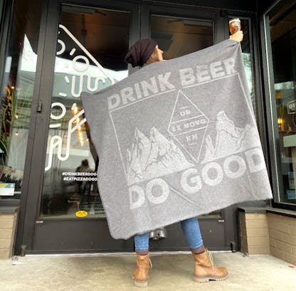 woman wearing black beanie and brown boots holds up beer and two tone gray blanket with mountains and large text "Drink Beer Do Good" and within diamond logo text "OR - Ex Novo - NM"