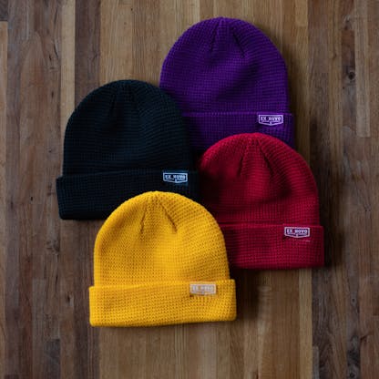 four waffle knit beanies purple, red, black and yellow overlaid on a wood table top each with a clip tag text "Ex Novo Brewing Company"
