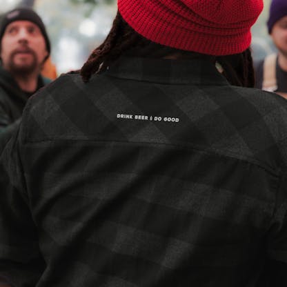 close up of back shoulders of man wearing red beanie and black and charcoal gray plaid flannel. white embroidered text "Drink Beer Do Good". in background two blurry faces can be seen.