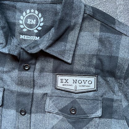 close up of black and charcoal flannel front collar, buttons, and left chest with pocket an custom chevron logo patch with text "Ex Novo brewing Company" inside collar features custom circular laurel logo with letters "E N" and word "MEDIUM"