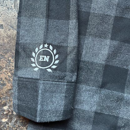close up of sleeve and cuff of black and charcoal plaid flannel with custom circular laurel logo and letters "EN" in center