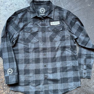 black and charcoal plaid button up flannel shirt on concrete floor with custom design elements: small circular laurel logo and letters "E N" on right cuff; internal printed circular laurel with "E N" inside color, text beneath "MEDIUM" and custom left front chevron patch above pocket text "Ex Novo"