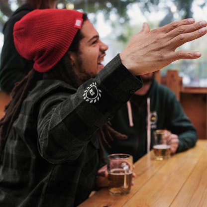 man sits at table with beer and friends right arm raised in gesture while talking. man wears black and charcoal gray plaid button up flannel with white embroidered cuff logo of circular laurel leaf and letters "E N", also wears red beanie with unreadable white text on clip tag. in background man wears green zip hoodie