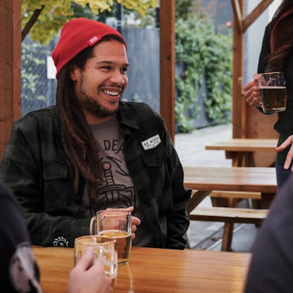 man sits at table with friends and beers wearing red beanie with unreadable clip tag, black and charcoal gray plaid flannel button up shirt with white chevron logo above left chest pocket text "Ex Novo". gray t-shirt partially visible with knife hilt image and partial text "DEA"