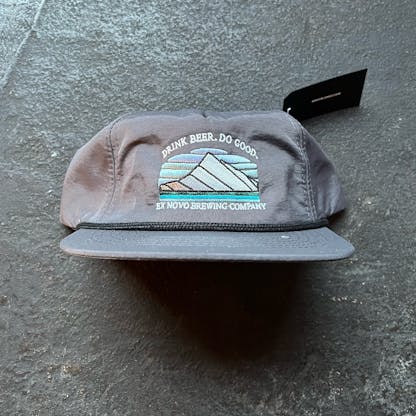 slate gray baseball hat rests on concrete floor with product tag up behind it, black rope runs along base of hat and bill, front features custom embroidery of a stylized mountain and text "Drink Beer. Do Good" arching over and "Ex Novo Brewing Company" beneath