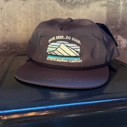 slate gray baseball hat sits atop a stool with concrete wall behind, black rope runs along base of hat and bill, front features custom embroidery of a stylized mountain and text "Drink Beer. Do Good" arching over and "Ex Novo Brewing Company" beneath