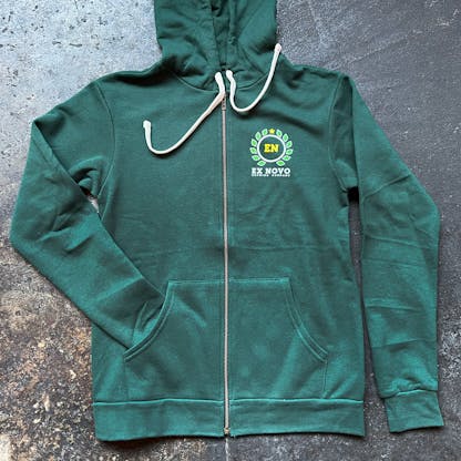 green zip up hoodie on concrete floor, front view fully zipped up, white strings flipped up, right arm tucked into pocket, left chest features yellow and light green circular laurel logo with yellow letters "E N" inside and white text beneath "Ex Novo Brewing Company"