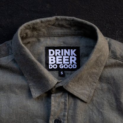 close up on collar of greyish sage green button up shirt custom black and white internal tag text "Drink Beer Do Good" and small size tag text letter "S"