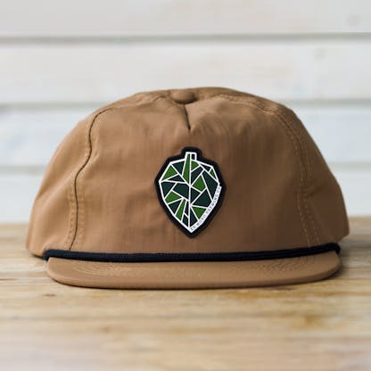 tan hat sits on tabletop, front with black rope along hat base and bill. pvc rubber patch of geometric hop cone or pinecone in green, dark green, and white with black outline. tiny text "Ex Novo Brewing Co"