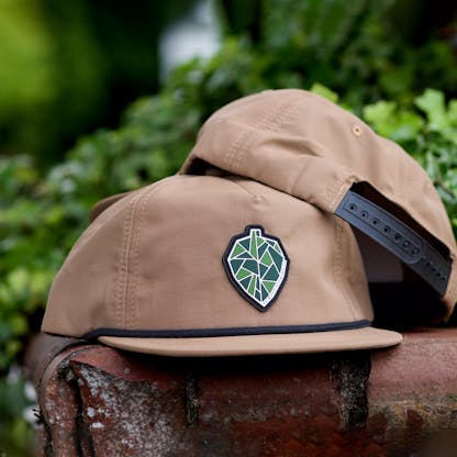 two tan hats stacked atop each other showing back black snapback closure and front with black rope along hat base and bill. pvc rubber patch of geometric hop cone or pinecone in green, dark green, and white with black outline. tiny unreadable text "Ex Novo Brewing Co"