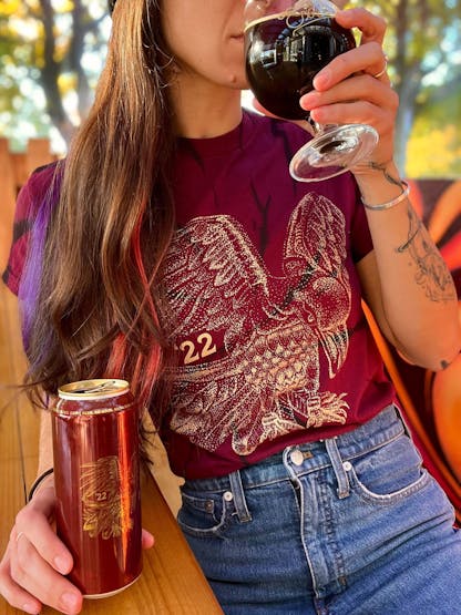 woman with long hair sits at a wood table, holds a maroon can with gold bird design and tiny number '22, wears a maroon shirt with same gold bird design and '22 print while drinking from a goblet of dark beer