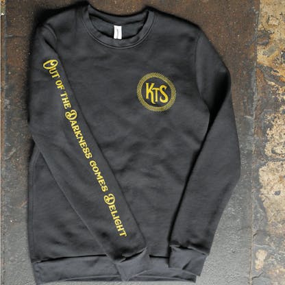black crewneck sweatshirt with gold text down the right arm reading "out of the darkness comes delight" and KTS small on the left chest