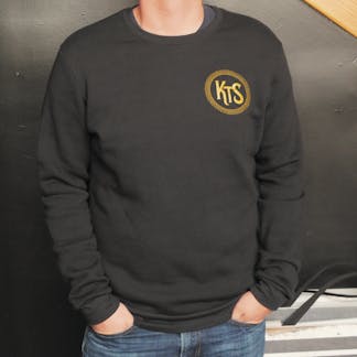 front view of person with hands in jean pockets wearing black crewneck sweatshirt with small circular gold logo containing letters "K T S"