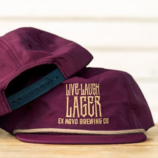 two maroon hats stacked atop each other showing back with black snapback closure and front with gold rope along hat base and bill, stacked gold lettering text "Live Laugh Lager Ex Novo Brewing Co"