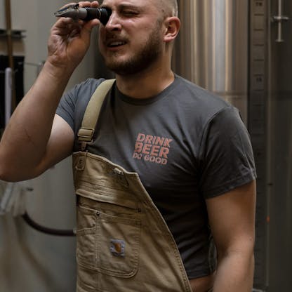 man looks through Brix meter device wearing gray crop top t-shirt with text "Drink Beer Do Good" and Carhartt overalls