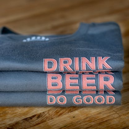 three gray shirts folded and stacked in a pile with pinkish-orange text "Drink Beer Do Good"