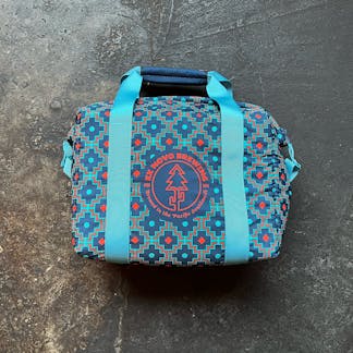 insulated cooler bag on concrete floor, dark orange, dark blue, and light blue geometric patterning, blue reinforced handle fabric, circular logo with text "Ex Novo Brewing" and "Brewed in the Pacific Southwest" surround stylized pine tree and cactus design