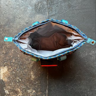 view down into insulated cooler bag on concrete floor. gray wrinkled interior, dark blue zipper, light blue handles partially visible and some geometric orange and two-tone blue pattern visible