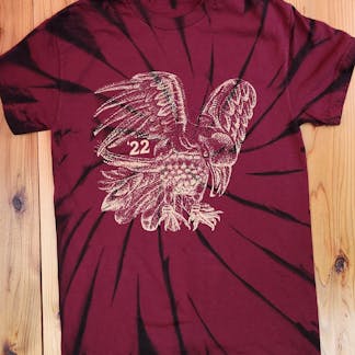 burgundy t-shirt with black swirl lines and gold printed bird design with number "'22"' on wood tabletop