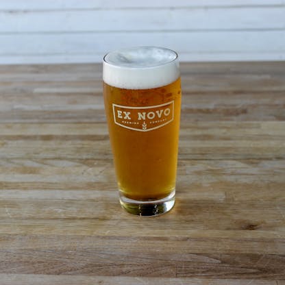 pint glass sits on tabletop filled with gold beer and thick white foam features gold colored chevron logo text" Ex Novo Brewing Company"