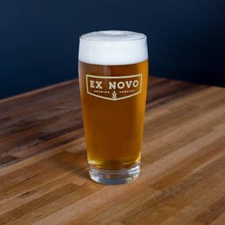 pint glass sits on tabletop filled with gold beer and thick white foam features gold colored chevron logo text" Ex Novo Brewing Company"
