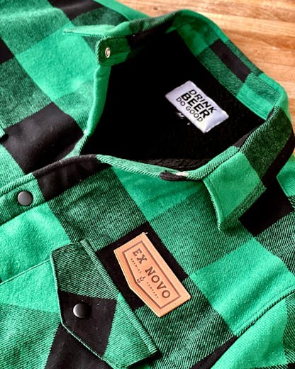 cropped close up of front collar of green and black plaid collared jacket with black fuzzy sherpa lining and black snaps. tan leather chevron logo patch above left chest pocket text "Ex Novo Brewing Company" and interior white tag with black stacked text "Drink Beer Do Good" above tiny size "M" tag