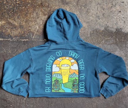turquoise cropped pullover hooded sweatshirt lays flat on concrete floor, back view shows bright multi colored beer glass in a valley setting with mountains and sunset in back and hills, a river, and evergreen tress in front. arched text around image reads "Ex Novo Brewing Co. Drink Beer Do Good" and center beer glass reads "Drink Lagers"