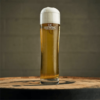 Tall slender pilsner glass filled with light lager sitting on top of a barrel.