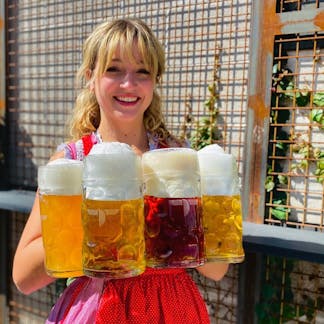 Female in Oktoberfest attire holding four liter mugs with different shades of beer.