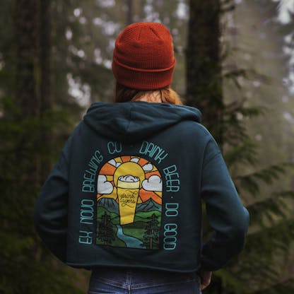 back view of female standing in foggy wooded setting, wearing rust orange beanie over red hair, hands in front pockets of blue jeans, wearing turquoise cropped pullover hooded sweatshirt, back view shows bright multi colored beer glass in a valley setting with mountains and sunset in back and hills, a river, and evergreen tress in front. arched text around image reads "Ex Novo Brewing Co. Drink Beer Do Good" and center beer glass reads "Drink Lagers"