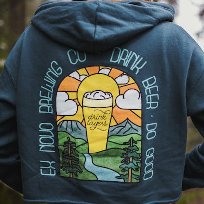 close up of female wearing turquoise cropped pullover hooded sweatshirt, back view shows bright multi colored beer glass in a valley setting with mountains and sunset in back and hills, a river, and evergreen tress in front. arched text around image reads "Ex Novo Brewing Co. Drink Beer Do Good" and center beer glass reads "Drink Lagers"