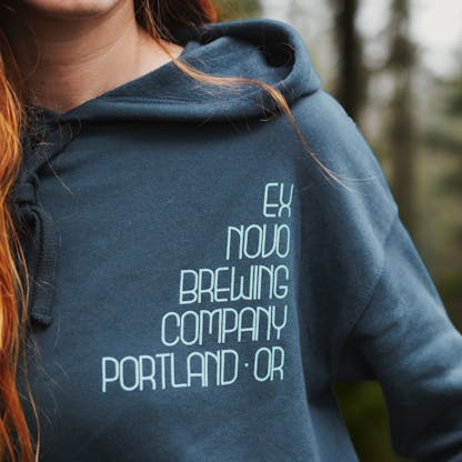 close up view of female wearing turquoise pullover hoodie, view of stacked text on front of hoodie that reads "Ex Novo Brewing Company Portland - OR"